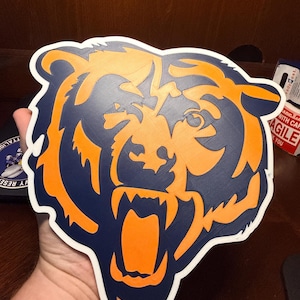 Könnte beinhalten: Zweifarbiges orange-blaues Bärenkopf-Logo mit weißem Rand. Der Bär knurrt, die Zähne sind entblößt. Das Logo ist wahrscheinlich für ein Sportteam oder eine ähnliche Organisation.
