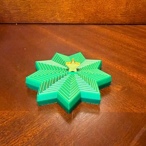 May include: A layered, star-shaped decorative object in shades of green and yellow. The design features a central star shape surrounded by concentric layers, creating a three-dimensional effect. The object is placed on a wooden surface.