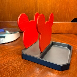 May include: A red and black desk organizer featuring a stylized mouse ear design. The organizer has a rectangular base with a divided tray and two upright red mouse ear shapes. The item is made of plastic.