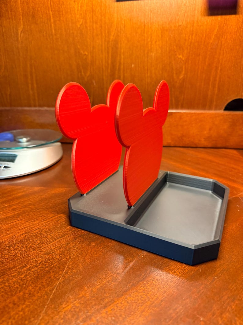May include: A red and black desk organizer featuring a stylized mouse ear design. The organizer has a rectangular base with a divided tray and two upright red mouse ear shapes. The item is made of plastic.