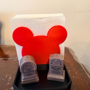 May include: A novelty kitchen set featuring a red Mickey Mouse-shaped napkin holder, two tombstone-shaped salt and pepper shakers, and a black tray. The salt and pepper shakers have humorous epitaphs. The napkin holder holds a stack of white paper napkins.