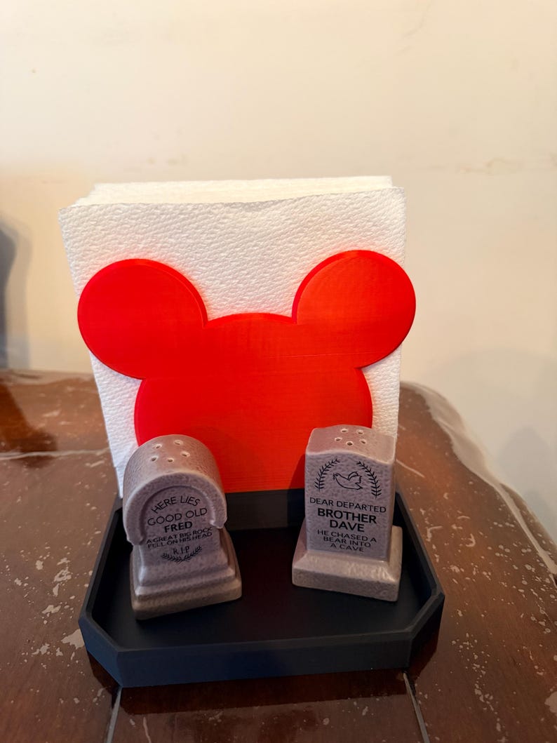 May include: A novelty kitchen set featuring a red Mickey Mouse-shaped napkin holder, two tombstone-shaped salt and pepper shakers, and a black tray. The salt and pepper shakers have humorous epitaphs. The napkin holder holds a stack of white paper napkins.