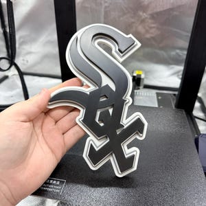 May include: A layered, three-dimensional logo of the Chicago White Sox baseball team. The logo features a stylized "S" and "X" in black and gray, outlined in white. The logo is held in a hand.