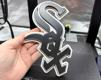 May include: A layered, three-dimensional logo of the Chicago White Sox baseball team. The logo features a stylized "S" and "X" in black and grey, outlined in white. The logo is held in a hand.