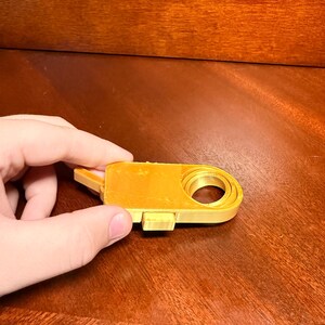 Può includere: Un oggetto giallo stampato in 3D con un corpo rettangolare e un'apertura circolare. L'oggetto ha una piccola linguetta sporgente su un lato. È tenuto da una mano su una superficie di legno.