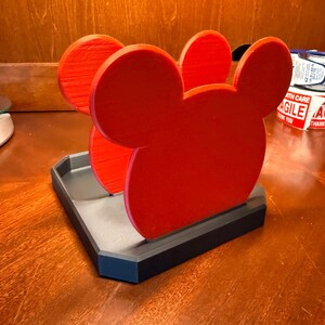 May include: A red Mickey Mouse-shaped desk organizer with a gray and black base. The organizer features three red Mickey Mouse head cutouts. The base is rectangular and has a raised edge.