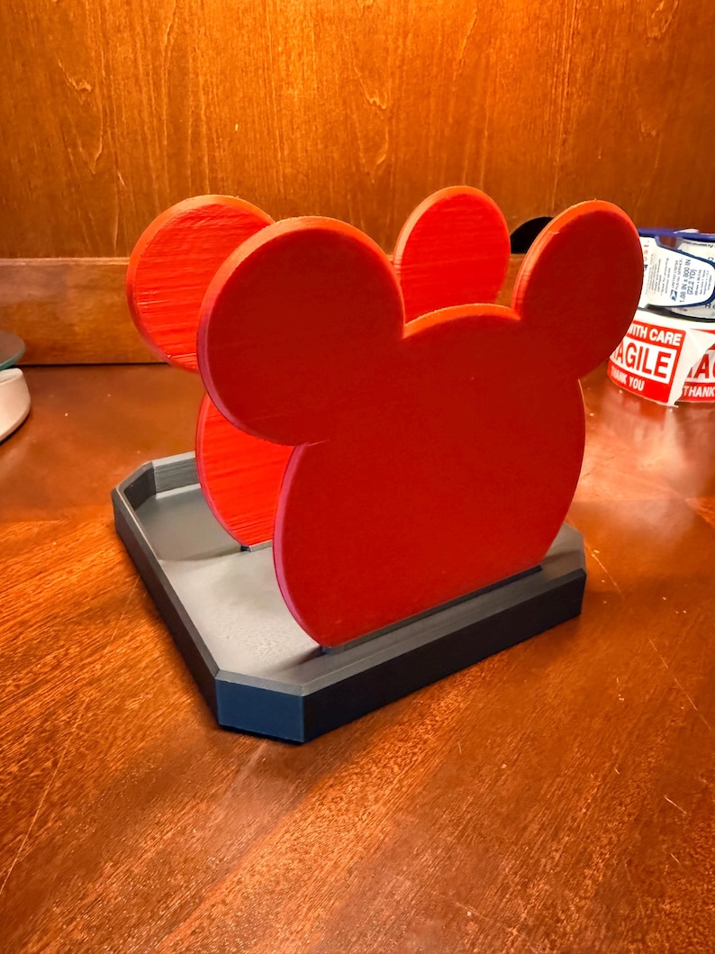 May include: A red Mickey Mouse-shaped desk organizer with a gray and black base. The organizer features three red Mickey Mouse head cutouts. The base is rectangular and has a raised edge.
