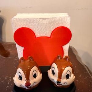 May include: A set of two Chip and Dale salt and pepper shakers with a red Mickey Mouse-shaped napkin holder. The shakers are brown and white with black eyes and red noses. The napkin holder is red and holds a stack of white napkins.