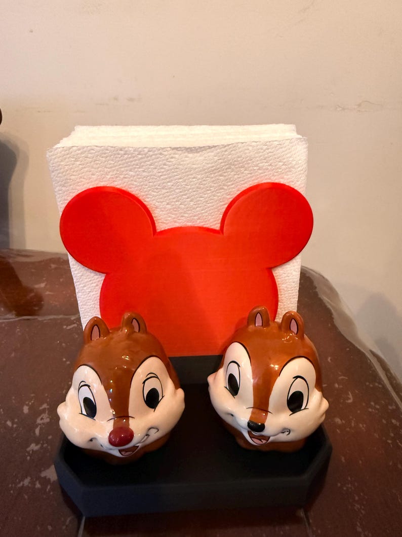 May include: A set of two Chip and Dale salt and pepper shakers with a red Mickey Mouse-shaped napkin holder. The shakers are brown and white with black eyes and red noses. The napkin holder is red and holds a stack of white napkins.