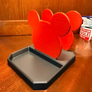 May include: A red Mickey Mouse-shaped desk organizer with a gray tray base and black trim. The organizer features three Mickey Mouse head silhouettes. The tray is rectangular with beveled edges.