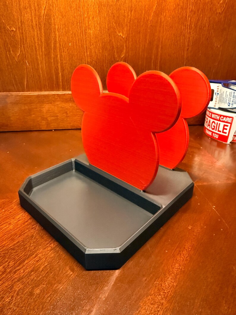 May include: A red Mickey Mouse-shaped desk organizer with a gray tray base and black trim. The organizer features three Mickey Mouse head silhouettes. The tray is rectangular with beveled edges.