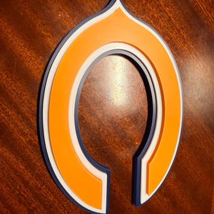May include: A layered, open-ended, decorative emblem in a stylized shape. The design features a vibrant orange center, bordered by a white layer, and outlined with a dark blue edge. The emblem is set against a dark wood grain surface.