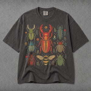 May include: A charcoal gray short-sleeve t-shirt featuring a central graphic print of various colorful beetles and a bee surrounded by small floral elements. The shirt has a crew neckline and a label at the inner collar that reads Comfort Colors.