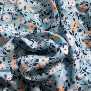 May include: Close-up of a light blue fabric with a floral pattern. The design features small white and peach-coloured flowers, along with dark blue leaves and stems. The fabric is gathered, creating soft folds and shadows.