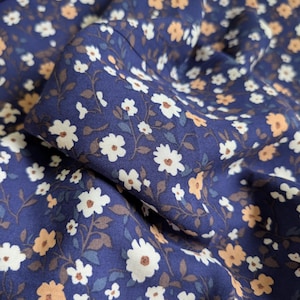 Può includere: Primo piano di un tessuto blu navy con motivo floreale. Il design presenta piccoli fiori bianchi e color pesca con steli marroni e foglie verdi. Il tessuto è piegato, mostrando la trama e i dettagli della stampa.