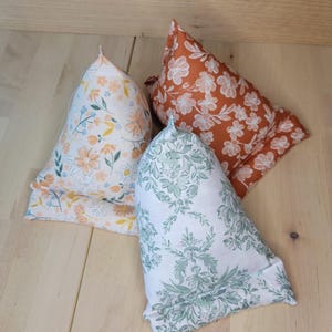 Puede incluir: Tres cojines decorativos para libros con estampados florales. Un cojín presenta un fondo blanco con flores naranjas y amarillas. Otro tiene un fondo color óxido con flores blancas. El tercer cojín tiene un fondo blanco con diseños florales verdes.