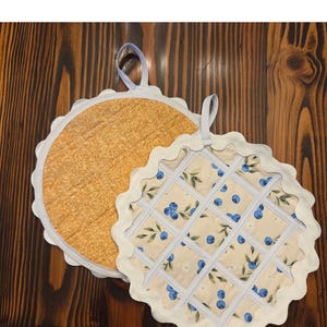 May include: Two round quilted pot holders with scalloped edges and fabric hanging loops. One pot holder features a golden-brown textured pattern, while the other displays a cream background with a blue blueberry and green leaf print overlaid with a white lattice.