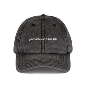 May include: A charcoal gray baseball cap with a curved brim. The cap features the white text "NEVER LET US GO" across the front. The hat has a vintage, worn look.