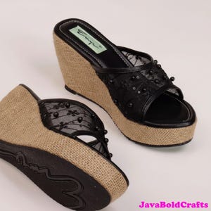 May include: Black wedge sandals with a jute-wrapped platform and heel. The sandals feature a black lace overlay adorned with small, dark beads. The footbed is black, and the brand name "Emye" is visible on the insole.