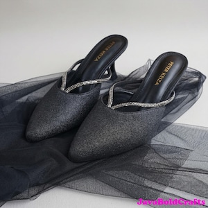 Könnte beinhalten: Ein Paar schwarze, glitzernde Mules mit Absatz, spitzer Zehenpartie und einem dünnen, mit Strasssteinen besetzten Riemen. Die Schuhe haben einen kleinen Absatz und der Markenname "PETER KEIZA" ist auf der Innensohle aufgedruckt.