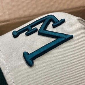 May include: A close-up of a baseball cap with a teal embroidered letter "M" on a light gray fabric. The cap has a structured design with a curved brim and a dark green underside.