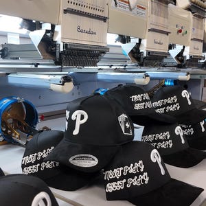 May include: Black baseball caps with white embroidered text and a "P" logo are stacked on a white surface. The caps have the text "BEST PLAY" and a playing card design. Industrial embroidery machines are in the background.