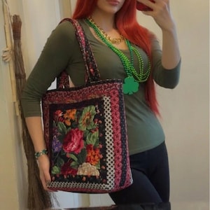 May include: A tote bag with a floral design featuring red roses and other colorful flowers. The bag has a black background with a patterned border. The bag is held in front of a mirror.