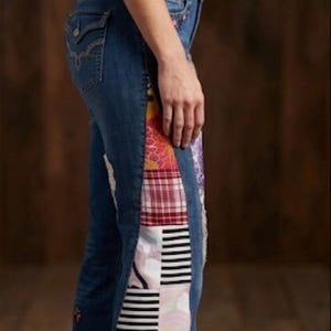 May include: Blue denim jeans featuring a vertical patchwork panel along the outer leg. The patchwork consists of various fabric patterns including red plaid, black and white stripes, floral prints, and geometric shapes. The jeans have a frayed hem.