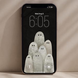 Pode incluir: Um smartphone exibindo um papel de parede digital com fundo preto e um grupo de seis figuras claras em forma de fantasma com olhos pretos simples. O texto Rose & Fern Printable PHONE WALLPAPER aparece acima da tela do dispositivo.