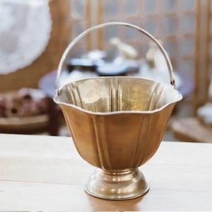 May include: A gold-colored metal decorative bowl with a fluted design and a handle. The bowl is sitting on a wooden surface.