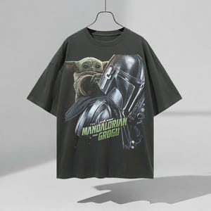 Pu&ograve; includere: T-shirt grigio scuro con una grafica di Grogu e il Mandaloriano di Star Wars. Il testo "THE MANDALORIAN AND GROGU" &egrave; stampato in verde. La maglietta &egrave; appesa a una gruccia nera.