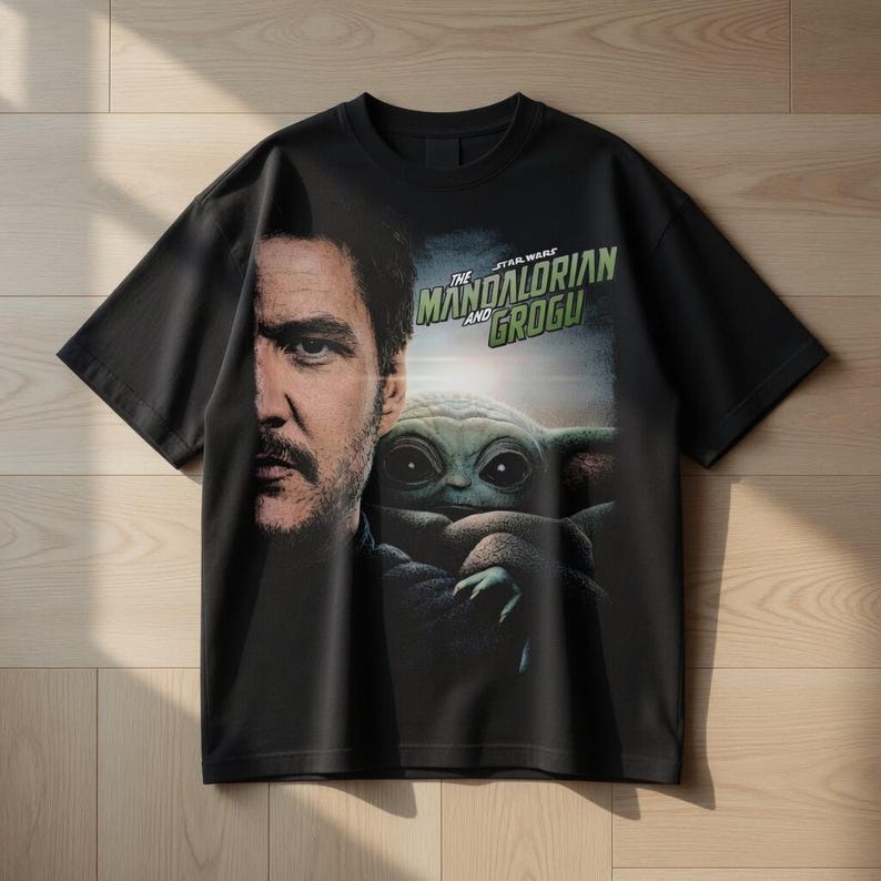 May include: Black t-shirt featuring a graphic of Pedro Pascal and Grogu from The Mandalorian. The Star Wars logo is printed in green and white. The t-shirt is made of a soft material and has short sleeves.