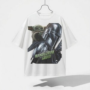 Pu&ograve; includere: T-shirt bianca con una grafica di The Mandalorian e Grogu di Star Wars. Il design include l'elmo del Mandaloriano e Grogu, con la scritta "The Star Wars Mandalorian and Grogu" in verde.
