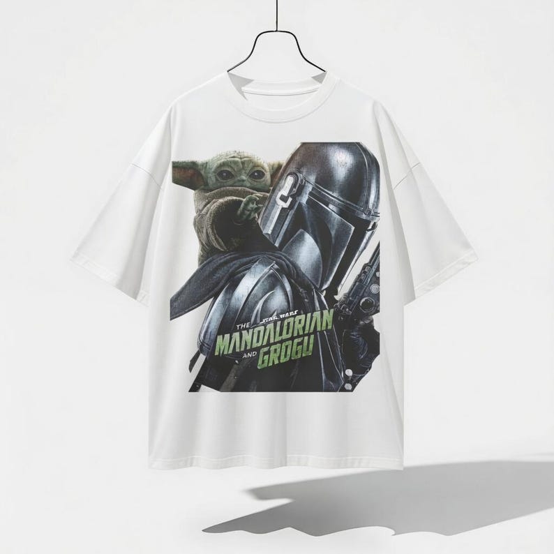 Pu&ograve; includere: T-shirt bianca con una grafica di The Mandalorian e Grogu di Star Wars. Il design include l'elmo del Mandaloriano e Grogu, con la scritta "The Star Wars Mandalorian and Grogu" in verde.