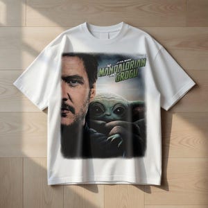 May include: White t-shirt featuring a graphic from the Star Wars series, "The Mandalorian and Grogu." The design includes a portrait of a man and a depiction of Grogu, also known as Baby Yoda. The text "The Mandalorian and Grogu" is in green.