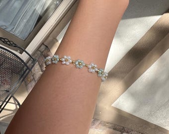 May include: A delicate bracelet featuring a floral design. Each flower is composed of white and light blue beads, with a gold-colored bead at the center. The bracelet is strung together with a thin, gold-colored chain, creating a charming accessory.