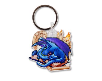 May include: A blue dragon keychain with purple wings, sleeping on an open book with a quill pen and candles. The keychain is attached to a silver ring.