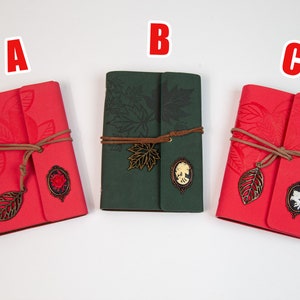 May include: Three small journals with decorative covers. The journals are red, green, and red. Each journal has a metal leaf charm and a metal oval charm with a design.