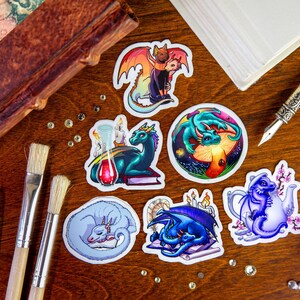 Peut inclure: Six autocollants color&eacute;s repr&eacute;sentant diff&eacute;rents dragons dans diverses poses. Les autocollants sont sur une surface en bois avec des pinceaux et un carnet.