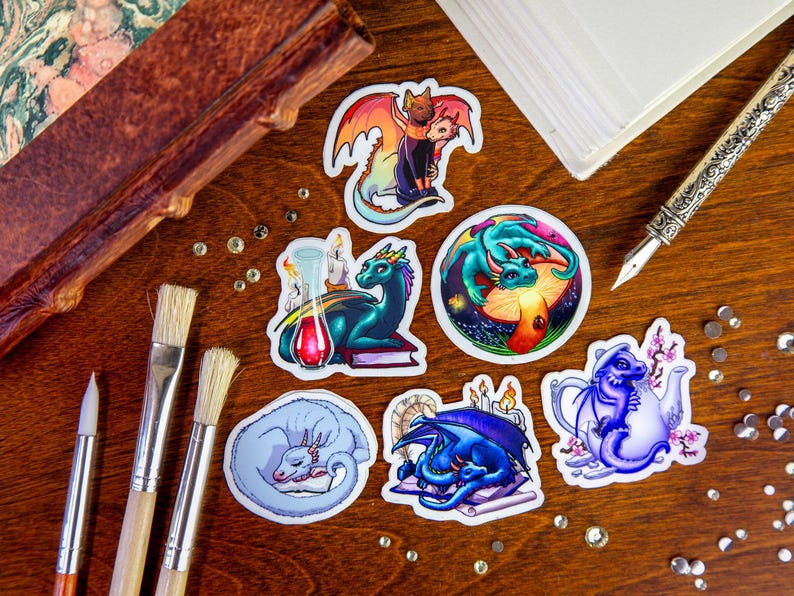 Peut inclure: Six autocollants color&eacute;s repr&eacute;sentant diff&eacute;rents dragons dans diverses poses. Les autocollants sont sur une surface en bois avec des pinceaux et un carnet.