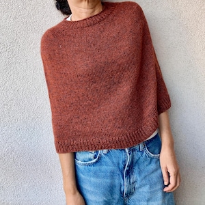 Puede incluir: Un suéter de punto color óxido con cuello redondo y mangas cortas. El suéter es de color liso y parece estar hecho de un hilo suave y texturizado. El modelo lleva vaqueros azules.