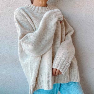 May include: Oversized cream-colored knit sweater with a ribbed neckline and cuffs. The sweater has long, voluminous sleeves and a relaxed fit. The soft, textured knitwear is a neutral tone, suitable for layering. A cozy, comfortable garment for cooler weather.