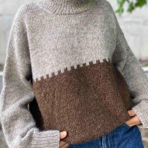 Könnte beinhalten: Ein zweifarbiger Pullover mit hohem Kragen. Die obere Hälfte ist hellgrau und die untere Hälfte ist ein sattes Braun. Die beiden Farben sind durch ein Zickzackmuster getrennt. Der Pullover hat lange Ärmel und eine lockere Passform.