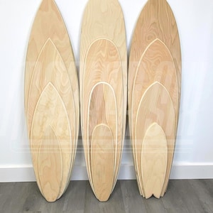 May include: Three wooden surfboard-shaped balance boards, each nested inside the other, with the largest board at the back and the smallest board at the front. The boards are made of light-colored wood with a natural grain pattern. The boards are arranged in a row on a gray wooden floor. The text "MARKER SIX" is visible in the lower left corner of the image.