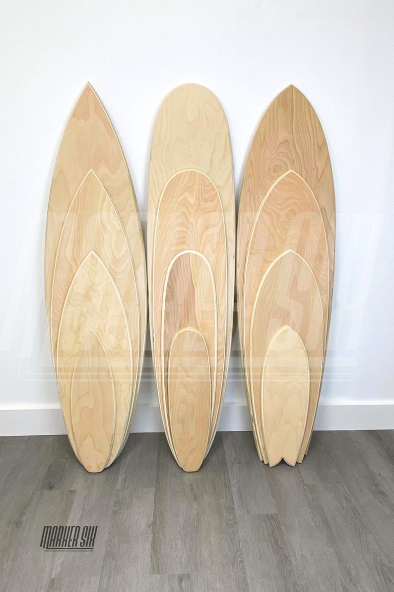 May include: Three wooden surfboard-shaped balance boards, each nested inside the other, with the largest board at the back and the smallest board at the front. The boards are made of light-colored wood with a natural grain pattern. The boards are arranged in a row on a gray wooden floor. The text "MARKER SIX" is visible in the lower left corner of the image.