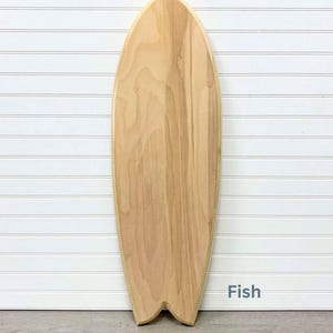 May include: A wooden surfboard shaped like a fish. The surfboard is light brown and has a natural wood grain. The surfboard is on a white background with a wood floor.