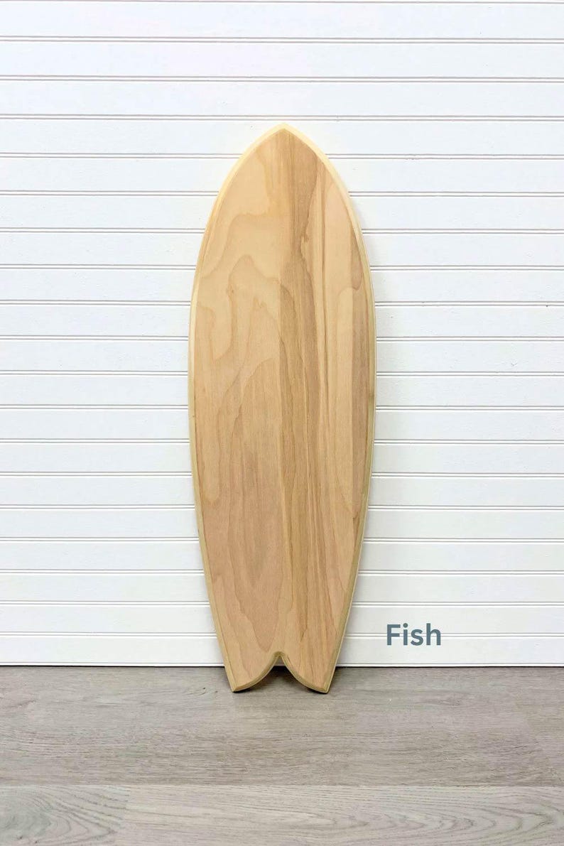 May include: A wooden surfboard shaped like a fish. The surfboard is light brown and has a natural wood grain. The surfboard is on a white background with a wood floor.