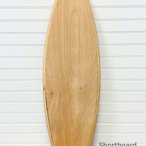 May include: A wooden surfboard, shaped like a shortboard, with a natural wood finish. The surfboard is leaning against a white wall with vertical wood paneling.