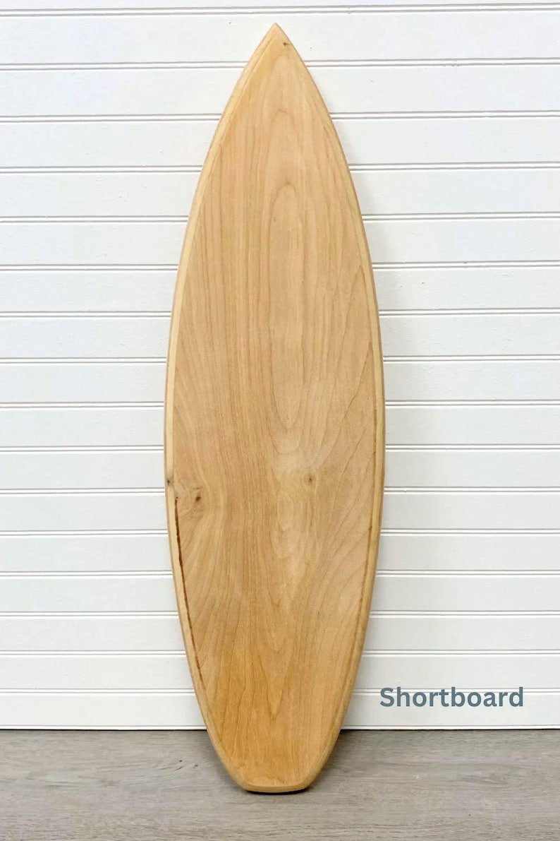 May include: A wooden surfboard, shaped like a shortboard, with a natural wood finish. The surfboard is leaning against a white wall with vertical wood paneling.