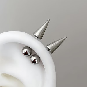 Op de afbeelding: Een zilverkleurige oorpiercing-set bestaande uit twee gladde, bolvormige knopjes en twee puntige, kegelvormige stekers. Het sieraad wordt getoond op een wit oormodel en heeft een metallic afwerking met een gepolijst, reflecterend oppervlak.